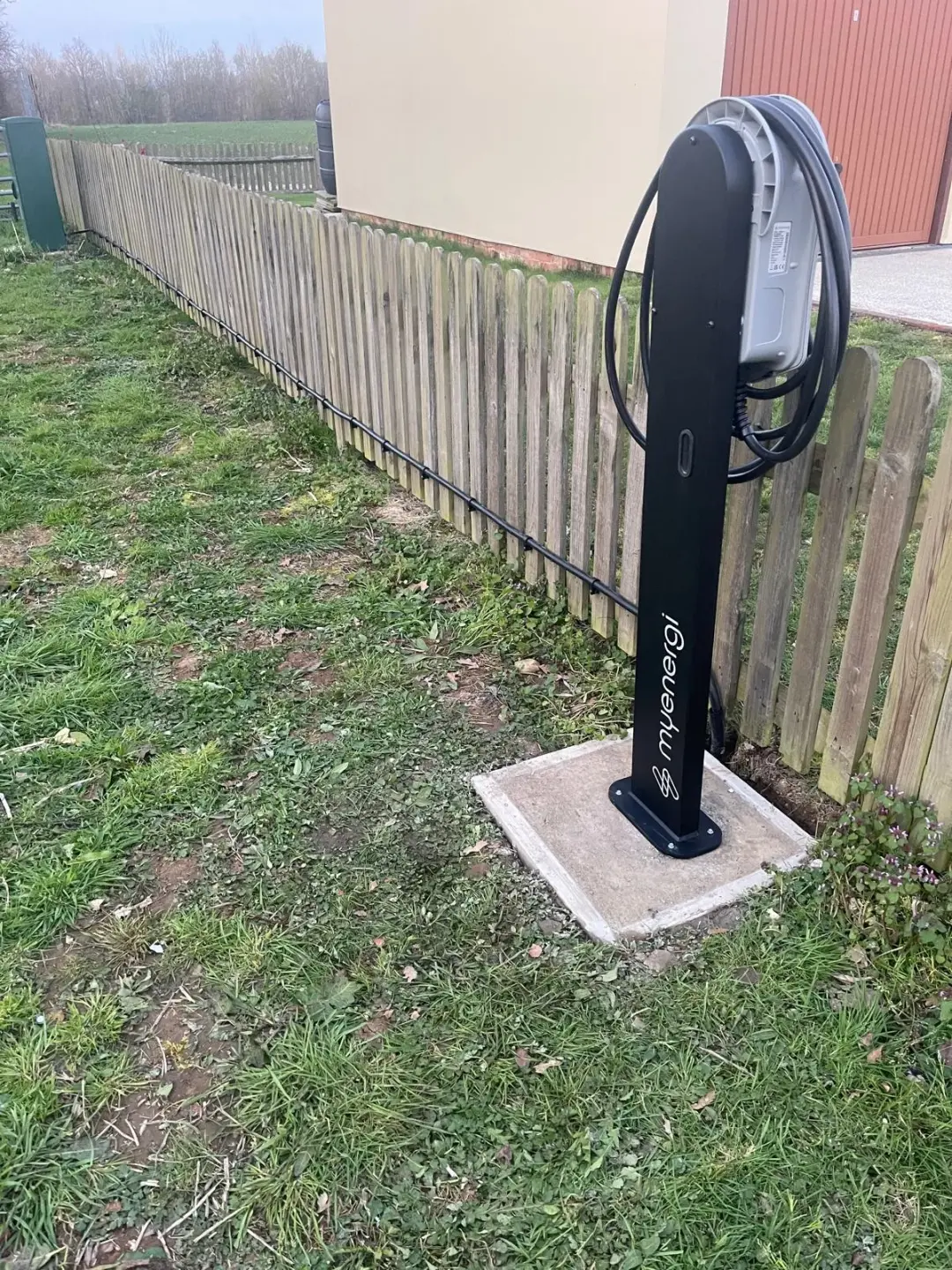 Pedestal-mounted EV charger with its supply cable routed along a wooden fence