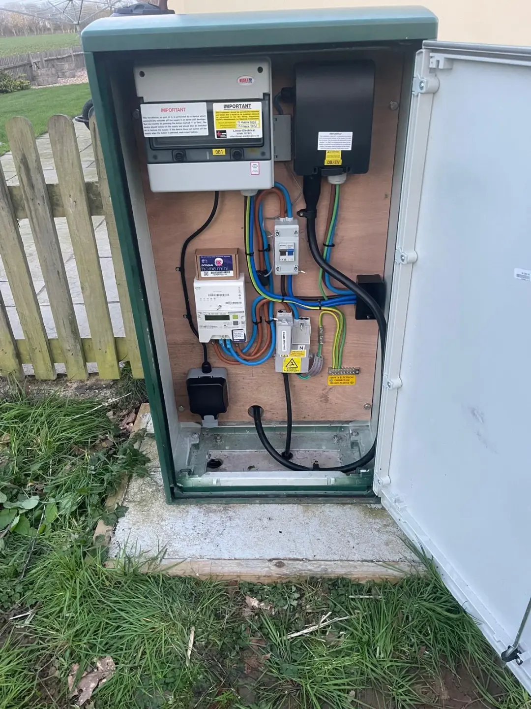 Open green outdoor electrical cabinet beside a picket fence with labelled distribution boards