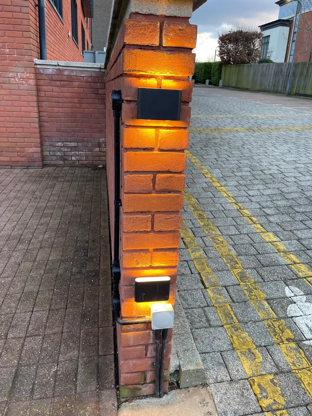 Warm exterior lights illuminating a brick entrance pillar beside a paved driveway