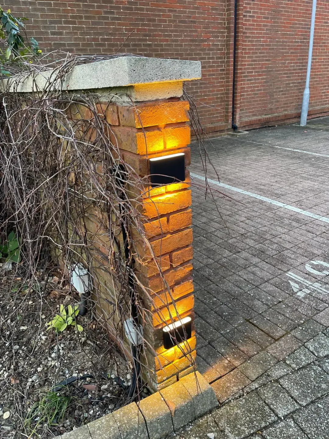 Two black outdoor lights casting warm light up and down a brick gate pillar